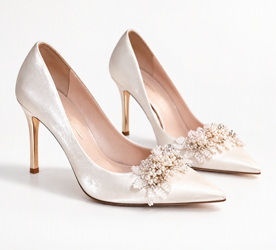 White pointed-toe bridal pumps with pearl-effect floral embellishments and gold-tone stiletto heels on a white background