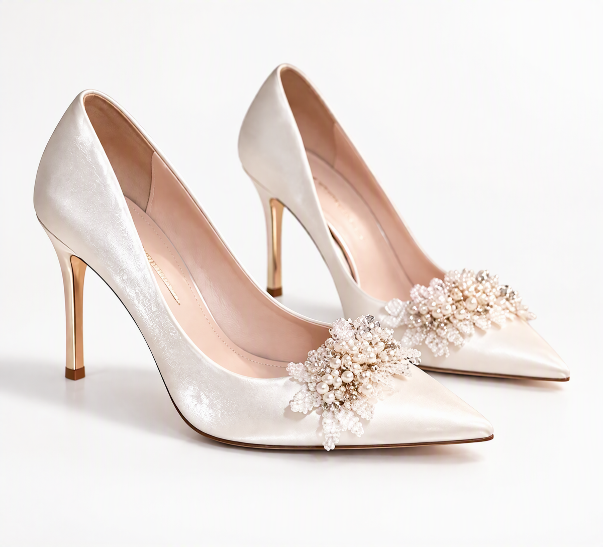 White pointed-toe bridal pumps with pearl-effect floral embellishments and gold-tone stiletto heels on a white background